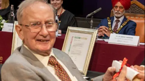 Austin appears to be sitting in a council meeting and is turning around to the camera. He has a grey suit on and white shirt