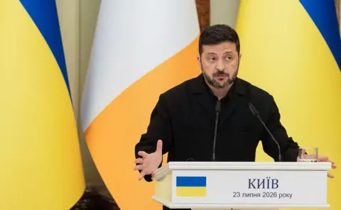 Tetiana DZHAFAROVA / AFP via Getty Images Zelensky looks to the right of frame while gesturing with his hands as he speaks from behind a lecturn with the Ukrainian flag on it, with Ukrainian and Irish flags in the background, in Kyiv on 23 July.