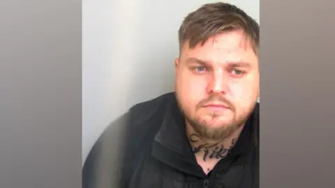 A mugshot of man with short light brown hair and a short beard. He has a tattoo on his neck.