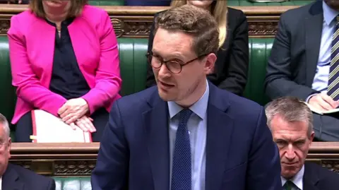 Chief Secretary to the Prime Minister, Darren Jones, makes a statement on the Lord Mandelson files in the House of Commons. He is wearing a navy suit, a blue shirt and a navy tie with tortoiseshell spectacles.