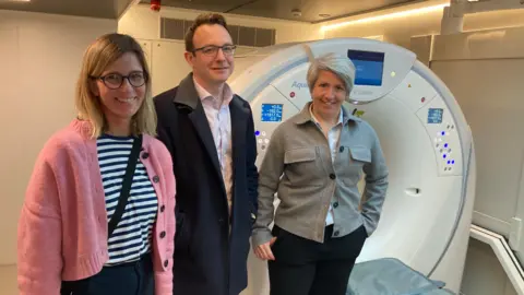 Dr Claudia Wegner, wearing a stripy top and pink cardigan, Dr Henry Steer, wearing a light dark jacket and white shirt and Dr Anna Bibby wearing a light shirt and grey jacket, all standing by a CT scanner. 