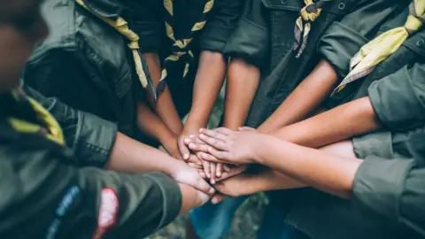 Group of young scouts, showing only their arms and and hands, as they join hands together in a show of unity. Their dark green uniform sleeves are rolled up to below their elbows.