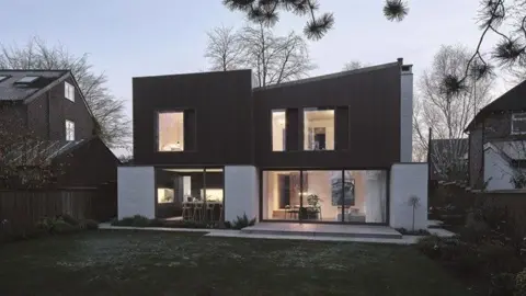 Riba A modern house that looks like two square building pushed together. The top half of the exterior is dark brown, the bottom half is made up of large windows. The building on the left has a flat roof while the building on the right has a slight diagonal slant.