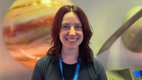 Heather Alexander is a young woman with shoulder-length, red hair. She is wearing a grey t-shirt and a blue lanyard and is smiling at the camera. In the background are some models of planets but they are slightly blurred.