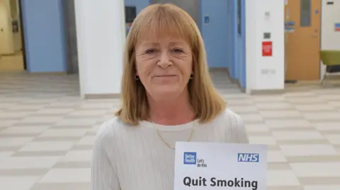 Royal Papworth Wendy Clark stands in a bright hallway in a hospital. She has blonde hair, she is wearing a white jumper and is holding a piece of paper that reads 'quit smoking'.