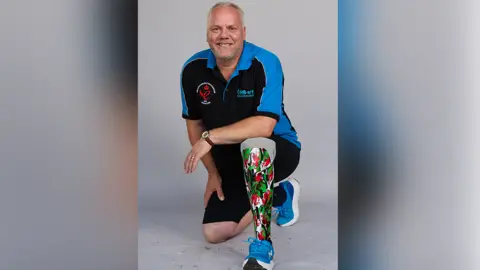 A middle-aged man wearing a blue and black t-shirt, with short receding white hair, kneels on the floor, and rests his elbow on the knee of his prosthetic leg. The prosthetic is in the green, white and red of the Welsh national flag.