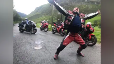 Handout A man wearing a red and white biker's jacket is standing up with his arms stretched out. There are four motorbikes behind him.