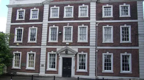 Andy Davis/Geograph The external front of the Grade II* listed Daresbury Hall. It is a three-store brick building with large white rectangle windows and a black door. It is very elegant and grand looking.