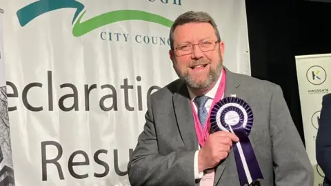 Chris Harper with short grey hair, a short beard and glasses. He is wearing a grey suit, white shirt and a blue tie, and is pointing to a round purple rosette pinned to his suit jacket following an election. He is smiling at the camera.