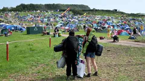 Getty Images Two people at Glastonbury Festival looking out over a camp site. There are lots of tents in the distance. The two people are standing with their backs to the camera. They are carrying luggage and camping equipment.