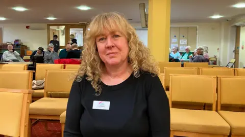 Liz has shoulder length curly blonde hair and wears a black top - she is sitting in front of rows of yellow coloured chairs with members of the music club in the background. 