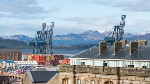 A yard full of shipping containers next to a river with snow-capped hills on the faraway bank