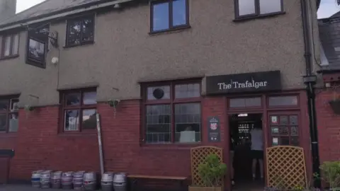 Outside view of a brick pub building with windows and a sign that reads “The Trafalgar,” with barrels lined up and a person entering the doorway.