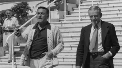 Richard Blanshard A black and white image of Gene Kelly and Fred Astaire walking down some steps in Cannes. Kelly is holding a pair of sunglasses and is wearing a dark polo shirt with a thick knit cardigan. Astaire is wearing a dark suit and tie. Both men have one hand in their pocket. 