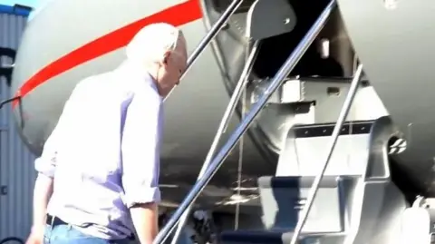 Julian Assange wearing light purple shirt and blue jeans walks up short flight of stairs into plane