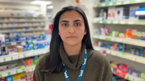 A woman with black hair stares directly into the camera. She has a dark green jumper on with a roll neck, and a blue lanyard with "well" printed on it. Behind her are shelves stacked with various medication.