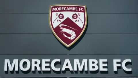 The Morecambe Football Club crest above the name 'Morecambe FC', which is written in capital letters, as seen on the front of their home ground, the Mazuma Mobile Stadium