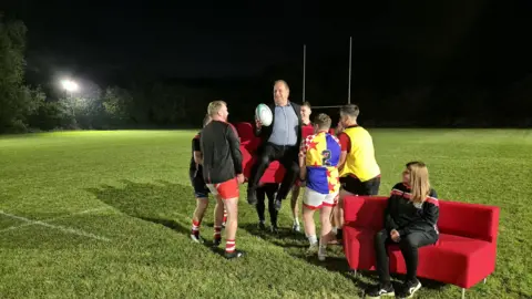 Rugby players at Claverdon RFC lift the red sofa, with reporter Ben Sidwell sitting on it, and carry it across the pitch