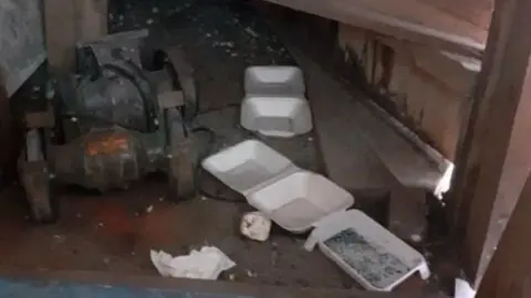 Kirklees Council Three white takeaway food containers on a dark, dirty floor. They have black specks inside them. A rusty piece of equipment in on the floor close to the containers. There is also a scrunched up paper napkin on the floor.