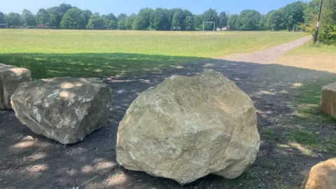 Boulders in front of a grassy playing field surrounded by trees on a bright sunny day.