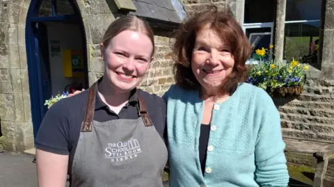 BBC/Seb Cheer A young woman and older woman stand next to each other, looking at the camera and smiling. The doorway to a former school building is in the background.