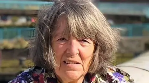 A woman with medium length grey hair and coat with flowers 