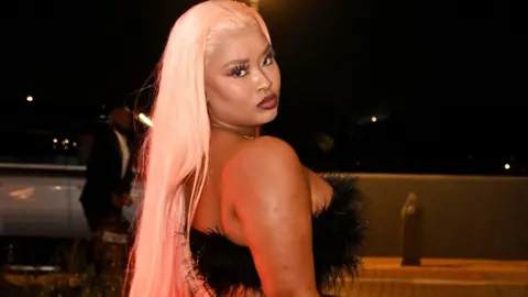 Cyan Boujee, with long blonde hair and wearing a black feathery bodice, looks over her shoulder as she poses for a photography at a hotel in Johannesburg in 2023.
