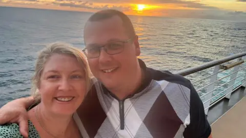 BBC Megan and Michael are standing on a boat with a sunset over the sea behind them. Michael has his arm around Megan's shoulder and they are smiling at the camera