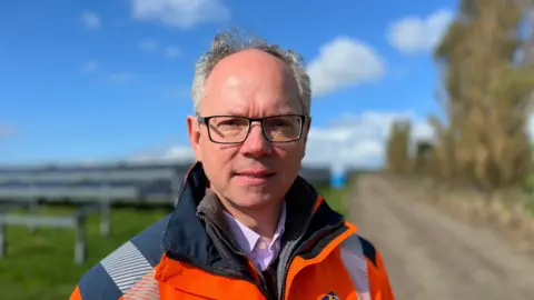 Chris Ambler has grey hair and wears glasses. He is wearing a a bright orange, high-viz coat. There are  solar panels in the background