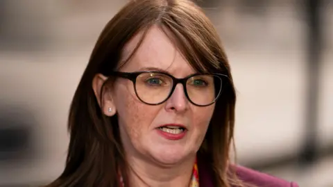 PA Media Caoimhe Archibald with brown/copper long hair and a side fringe. She is wearing dark framed glasses, a diamond stud earring in the ear not covered by her hair. She is mid-speech outside the Treasury in London. She is wearing a pink jacket and pink, red, yellow and orange floral scarf.