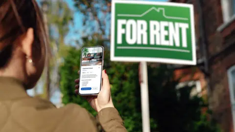 A woman in front of a green sign which says 'For Rent' in white letters. She is looking for information about the house on her phone and is wearing a beige jacket and has brown hair tied back. 