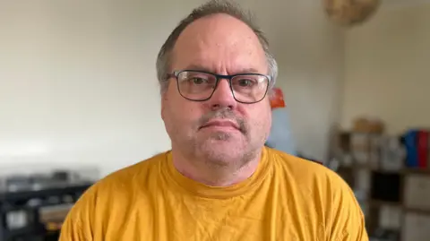 BBC A man in his fifties is looking into the camera with a neutral expression. He is sitting in his wheelchair in the lounge of his home. He is wearing a yellow t-shirt and has glasses on. 