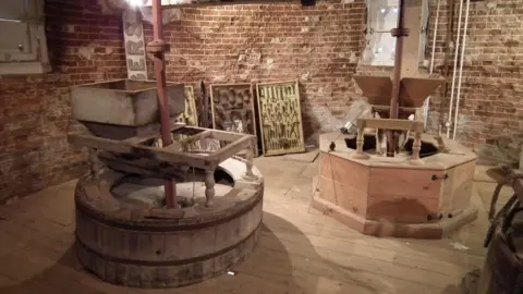 Trustees of Thaxted Windmill Inside the windmill, which has a brick wall and a wooden floor. There is various milling equipment