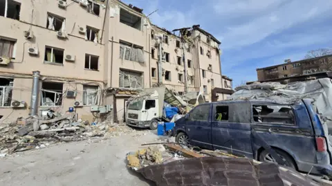 Karol Swiacki Some of the aftermath captured by Karol in daylight. Shows a beige building hit by the missile. All the glass from the windows is gone and there are sections of the roof missing and rubble covering the floor. There is a lorry filled with rubble and a blue van to the right of the image which has broken windows and another van behind it covered in rubble and covered in dents from the impact.