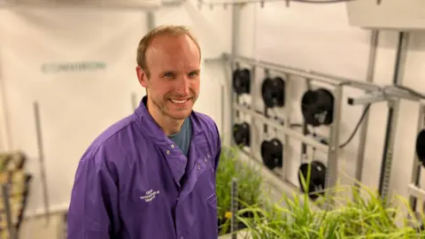 A man in a purple lab coat stands in a small lab, there are rice plants on silver shelves in the background and fans on the walls that simulate wind.