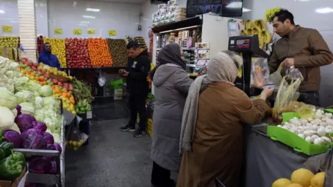 Scene inside a fruit and vegetable store, where two women are buying an item from a man at the till and two other men can be seen in the background.