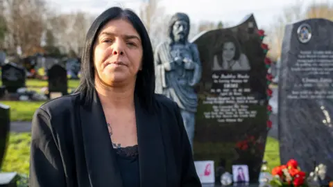 Helen se para frente a la tumba de su hija. Está vestida de negro y mira la cámara. 