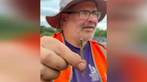 Uncovering Roman Carlisle Site director Frank Giecco holds a bone hairpin found during the dig at the cricket club in Carlisle. Mr Giecco is wearing a hat, glasses and a high-viz vest.
