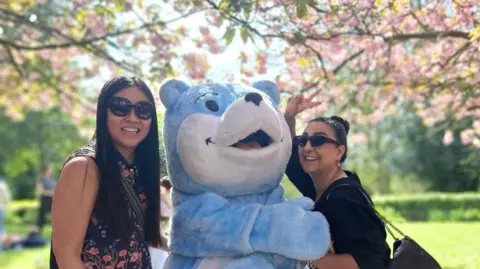 Bradford Council Two people stand outdoors beside a large blue‑and‑white bear mascot, all smiling beneath blooming pink cherry blossoms on a bright spring day.