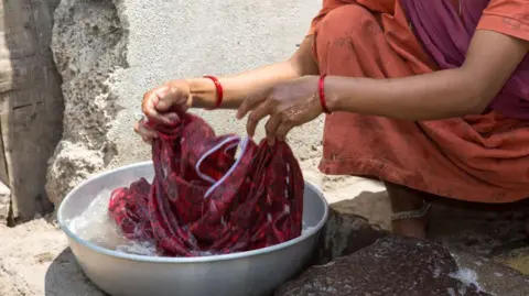 Getty Images Bijapur, India - 28 Maret: Seorang wanita dengan sari merah sedang mencuci kain di wajan cuci saat dia duduk di squat di depan gubuk pada 28 Maret 2013 di Bijapur, India. Sekitar 50.000 orang tinggal di daerah kumuh yang membentuk sekitar seperenam dari populasi Bijapur. (Foto oleh Christian Ender/Getty Images)