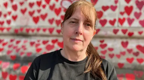 A woman with long brown hair worn in a ponytail looks into the camera with a thoughtful smile on her face. She stands in front of a wall with many red hearts painted onto it