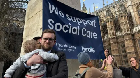 Freddie van Mierlo Freddie van Mierlo holding a child and standing in front of a sign saying, "We support Social Care".