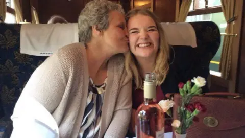 Jordan Findlay A mum and teenage daughter are pictured on a train. It looks like a luxury trip as there is a bottle of wine, glasses and napkins in front of them. The mum kisses the daughter's cheek and the daughter looks into the camera with a big smile.