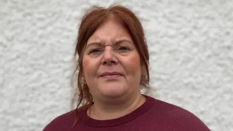 The image shows Michelle Brace, a woman with red hair and a burgundy top standing with her back to a white pebble-dash wall.
