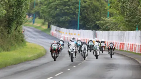 A group of motorcycles riding up the course at Billown, with crash barriers and fencing to the right and greenery to the left.