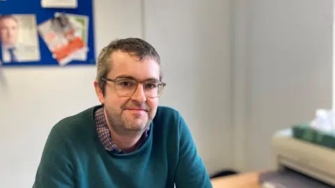 Laura Coffey/BBC Paul has short hair and glasses sitting at a desk. He is looking at the camera. He is wearing a spotty shirt and dark green jumper. In the background is a board with leaflets and to his left is a printer.