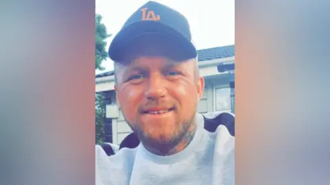 A photo of Marcus Carpenter. He is smiling at the camera, he has a beard and tattoos on his neck. He is wearing a grey sweatshirt with black details, and a black cap.