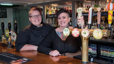 Star Pubs Two women stand behind a well stocked bar. They are both wearing black and smiling and one has a very colourful tattoo visible on her chest and neck. 