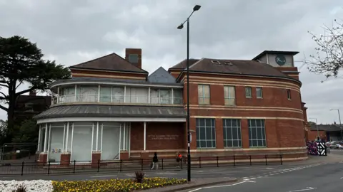 A large building on the corner of a town street with large windows that are covered with grey paint. Part of the building is red brick and has blacked out windows with blur frames. There is a small tower with a circular logo with the letters "BHS" on it