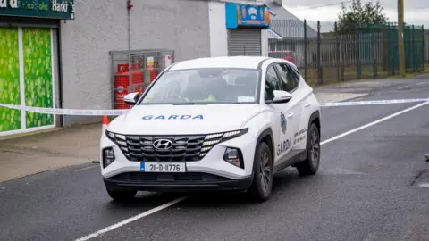 A garda car is parked on an empty road. Police tape is up. 